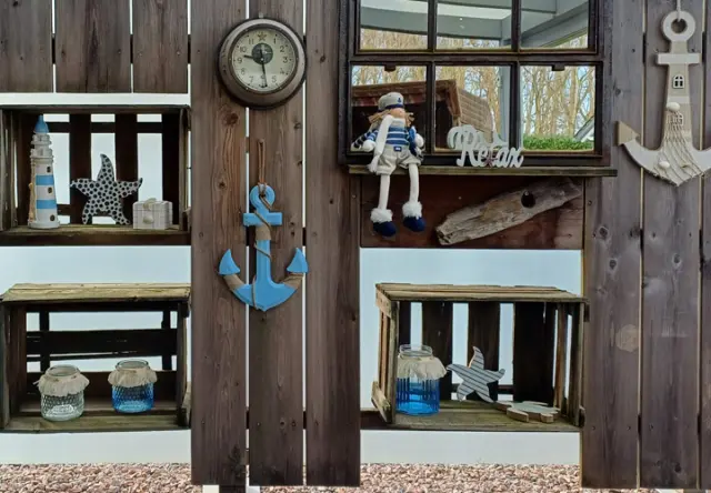 Urlaubsfeeling für jeden Tag Eine rustikale Holzwand zeigt nautisches Dekor, darunter eine Uhr, einen Anker, Regale, Seesterne und eine Matrosenpuppe vor einer gemütlichen Kulisse.