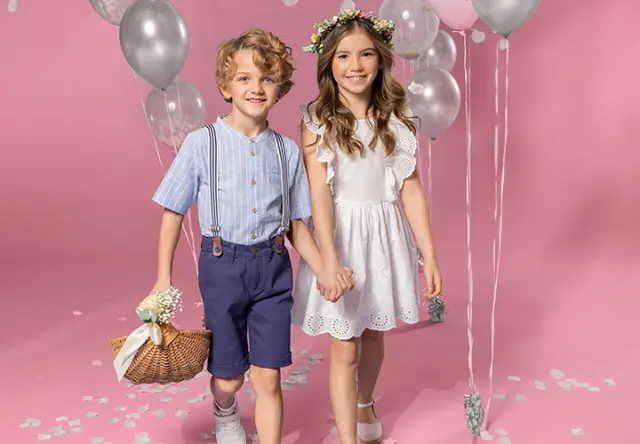 Sommerlooks für kleine Entdecker Zwei Kinder halten sich an den Händen. Die Kinder tragen festliche Klamotten und der Hintergrund ist rosa