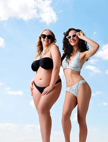 Zwei Frauen posieren selbstbewusst in Bikinis vor einem strahlend blauen Himmel und zelebrieren Körperbewusstsein und Sommerstimmung.
